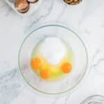 A glass bowl with three eggs and sugar, a whisk, a small bowl of walnuts, and cracked eggshells on a marble countertop.