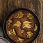 A round chocolate cake topped with sliced pears in a black baking pan on a wooden surface, with a beige cloth nearby.
