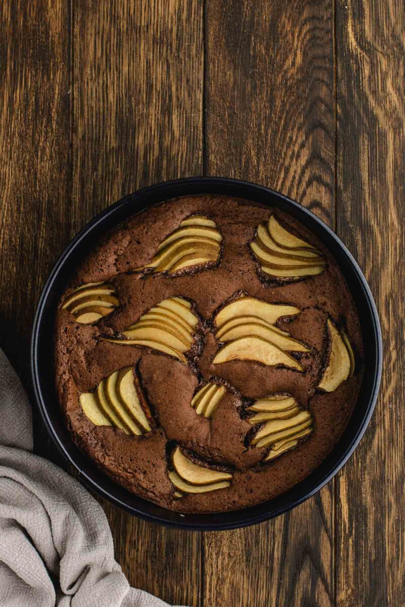 A round chocolate cake topped with sliced pears in a black baking pan on a wooden surface, with a beige cloth nearby.
