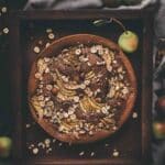 A round cake topped with sliced pears, oats, and hazelnuts sits on a dark tray, surrounded by fresh pears and a gray cloth.