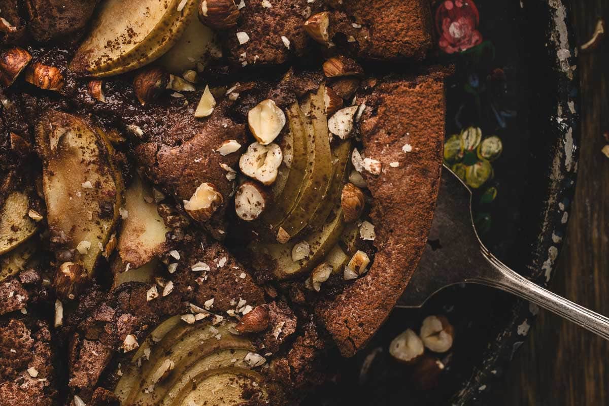 Close-up of a chocolate hazelnut cake topped with sliced pears and chopped hazelnuts, with a cake server lifting one slice.