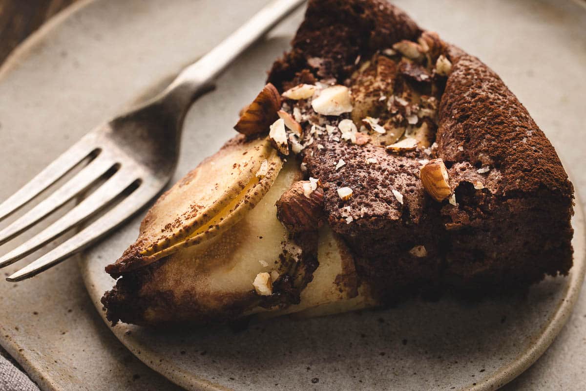 A slice of chocolate and pear cake topped with chopped hazelnuts on a ceramic plate with a fork.