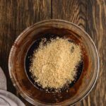 A glass bowl containing a mixture of ground nuts and chocolate batter sits on a dark wooden surface next to a gray cloth.
