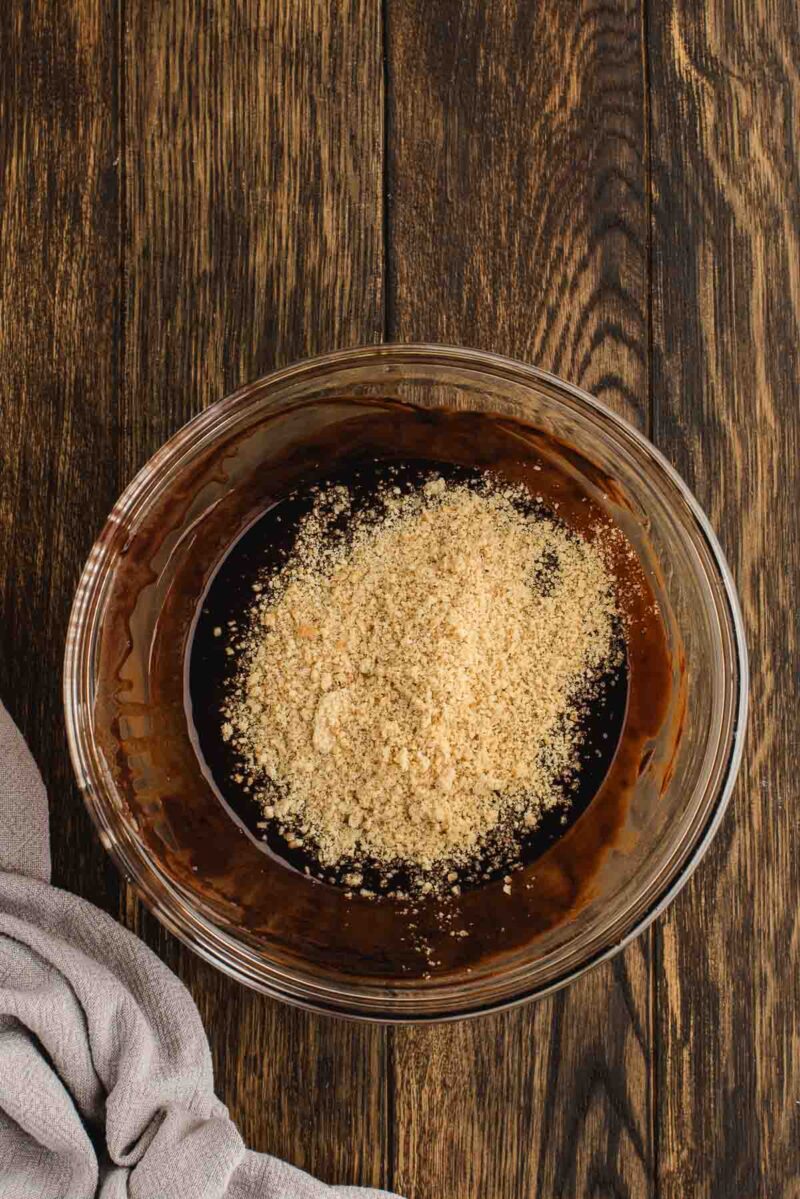 A glass bowl containing a mixture of ground nuts and chocolate batter sits on a dark wooden surface next to a gray cloth.