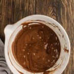 A white mixing bowl filled with chocolate cake batter sits on a dark wooden surface next to a gray cloth.