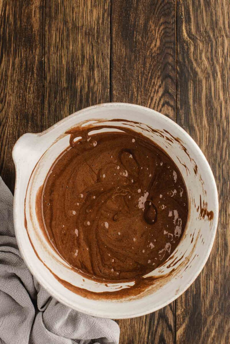 A white mixing bowl filled with chocolate cake batter sits on a dark wooden surface next to a gray cloth.