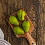 Three green figs are placed on a small wooden cutting board, set on a dark wooden surface next to a light gray cloth.