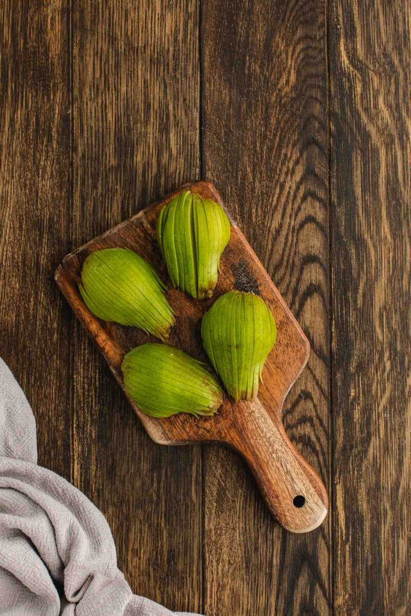Three green figs are placed on a small wooden cutting board, set on a dark wooden surface next to a light gray cloth.