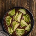 A round black dish filled with chocolate batter topped with thinly sliced green apples, placed on a dark wooden surface next to a beige cloth.
