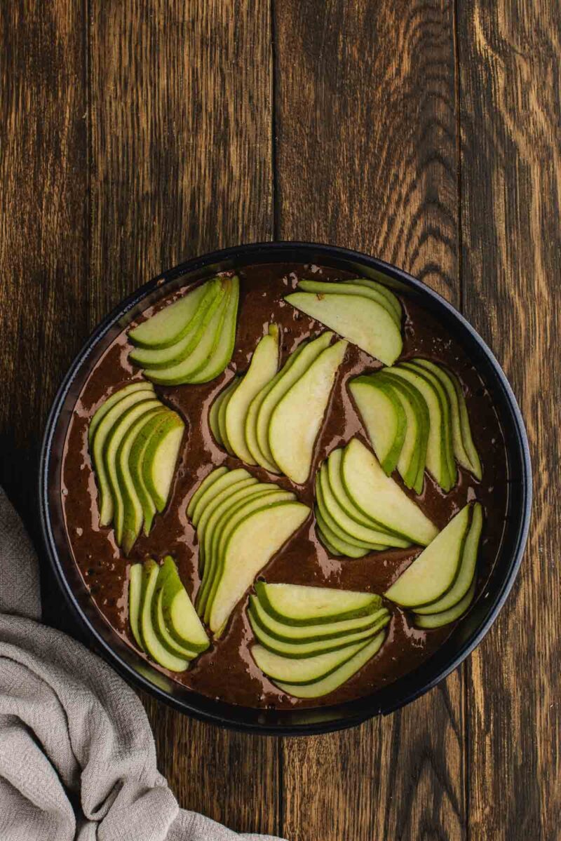 A round black dish filled with chocolate batter topped with thinly sliced green apples, placed on a dark wooden surface next to a beige cloth.