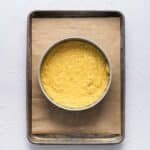 A round cake pan filled with yellow cake batter sits on a parchment-lined baking sheet against a white background.