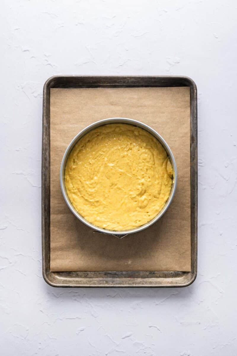 A round cake pan filled with yellow cake batter sits on a parchment-lined baking sheet against a white background.