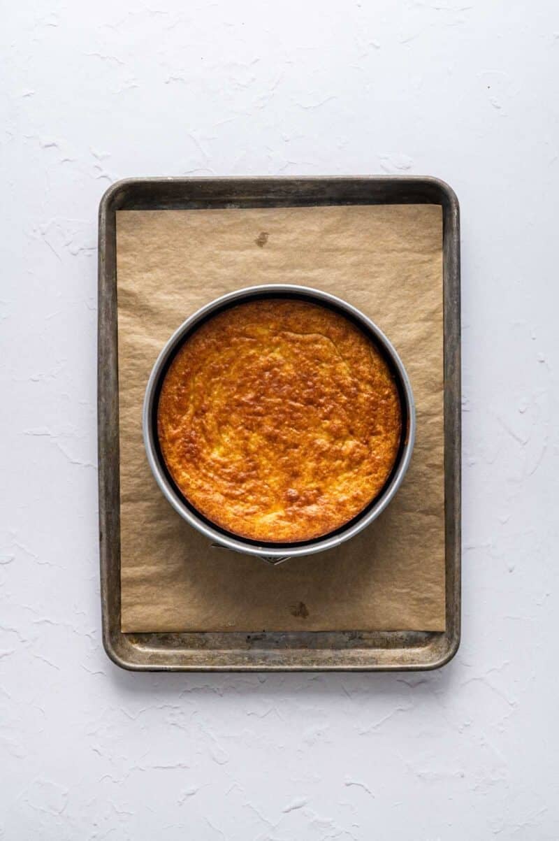 A round cake in a metal pan sits on a sheet of parchment paper on a baking tray, viewed from above on a white surface.