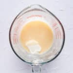 A glass measuring cup containing a light-colored liquid mixture with a dollop of sour cream on top, placed on a light textured surface.