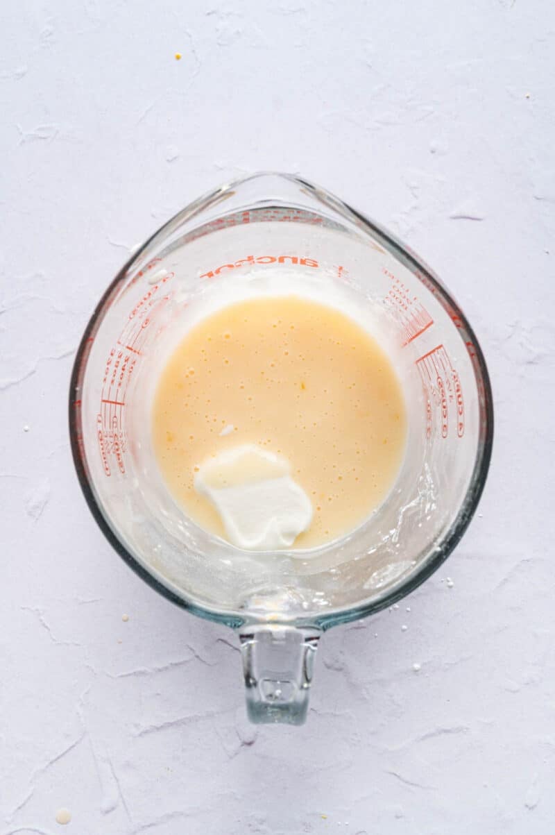 A glass measuring cup containing a light-colored liquid mixture with a dollop of sour cream on top, placed on a light textured surface.