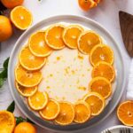 A round clementine cake topped with a ring of orange slices sits on a white plate, surrounded by whole and cut oranges, a glass of orange juice, and plates with forks.