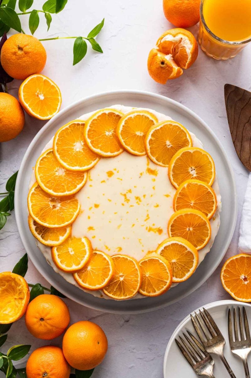 A round clementine cake topped with a ring of orange slices sits on a white plate, surrounded by whole and cut oranges, a glass of orange juice, and plates with forks.