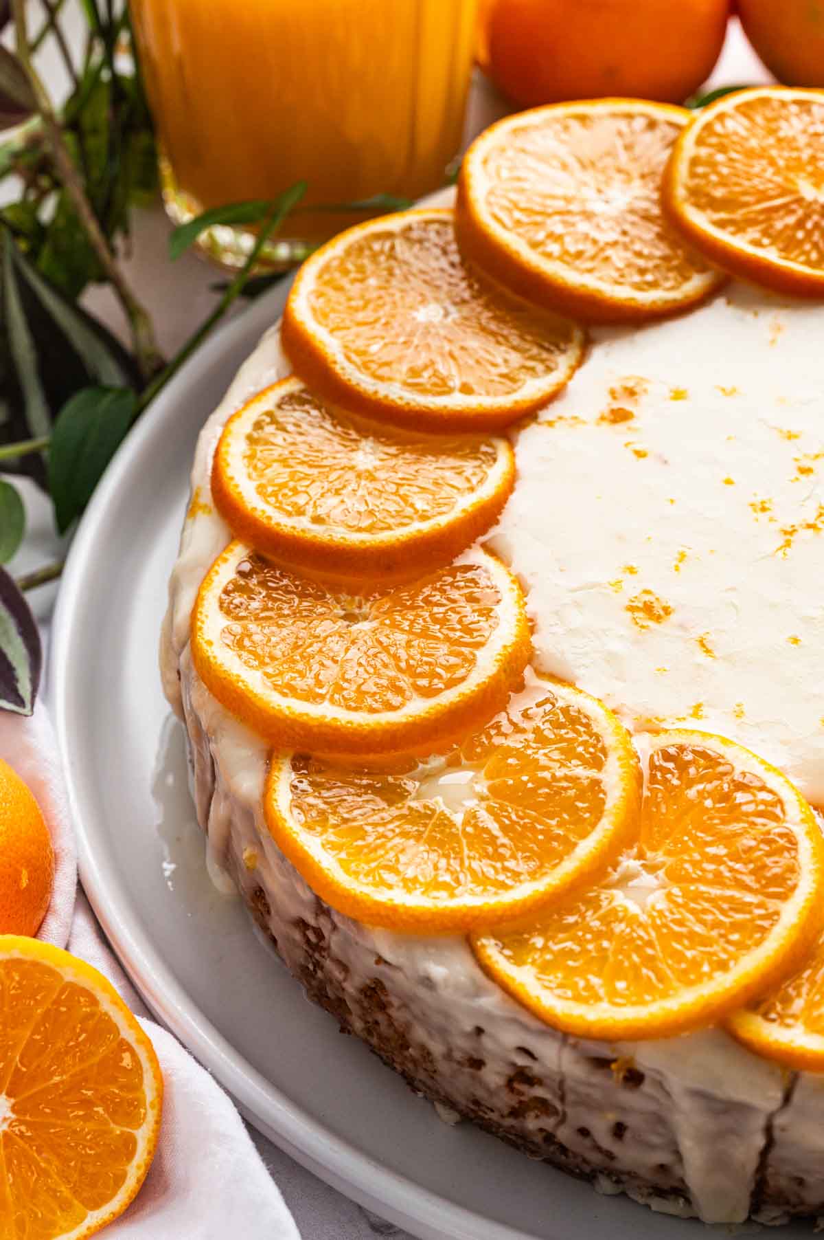A round clementine cake with white icing, decorated with overlapping orange slices around the edge, sits on a white plate.