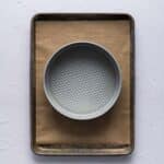 A round metal springform pan sits on a sheet of brown parchment paper placed on a rectangular baking tray.