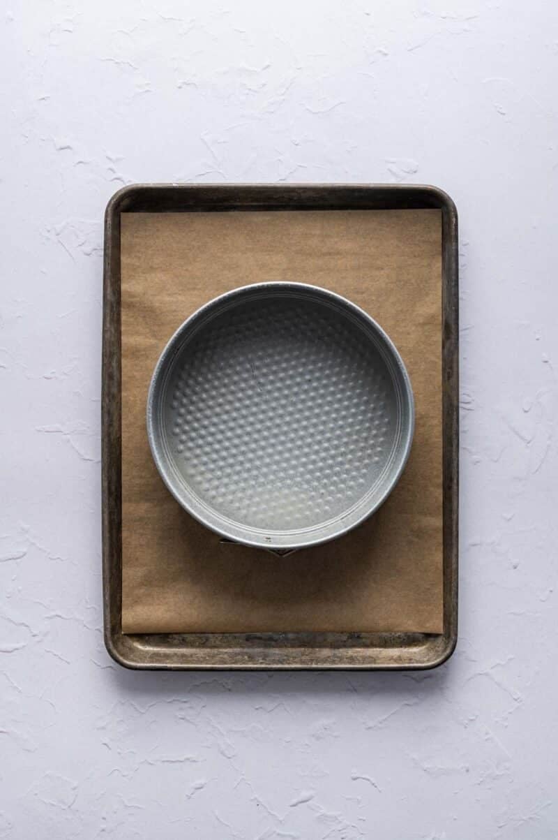 A round metal springform pan sits on a sheet of brown parchment paper placed on a rectangular baking tray.