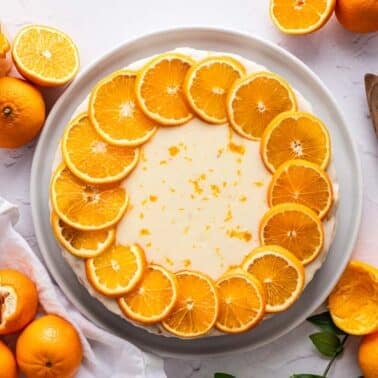 A round clementine cake topped with a ring of orange slices, surrounded by whole and halved oranges, a glass of orange juice, and a wooden serving utensil on a white surface.