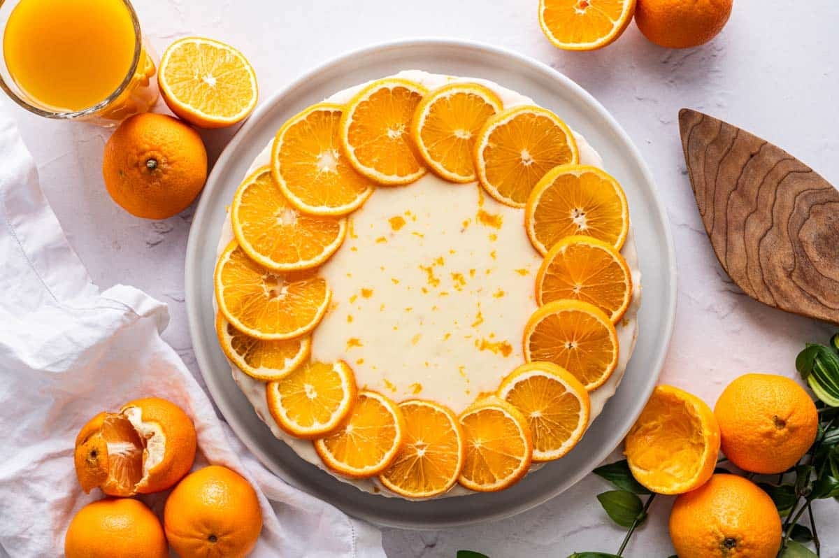 A round clementine cake topped with a ring of orange slices, surrounded by whole and halved oranges, a glass of orange juice, and a wooden serving utensil on a white surface.