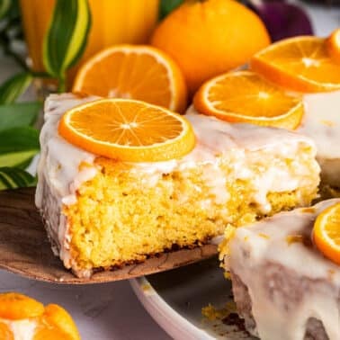 A slice of orange clementine cake with white icing and orange slices on top is being lifted from a whole cake on a plate.