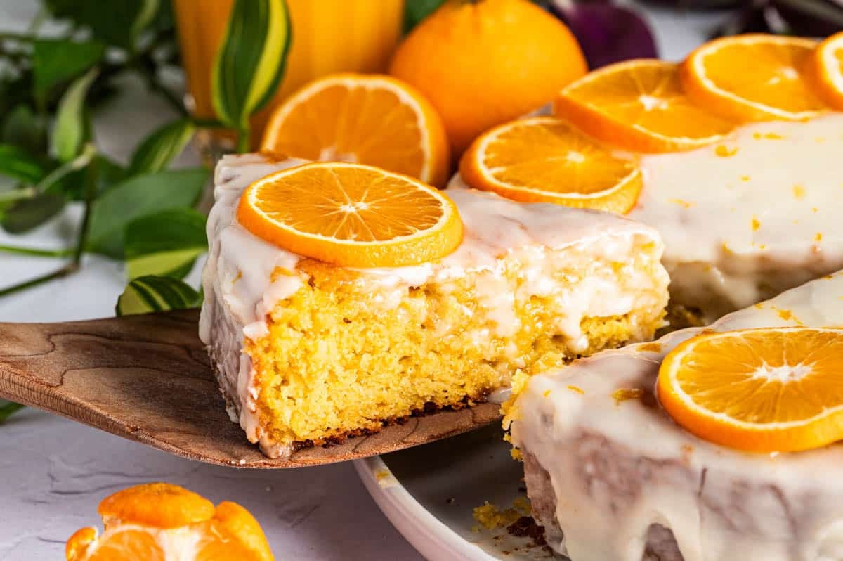A slice of orange clementine cake with white icing and orange slices on top is being lifted from a whole cake on a plate.