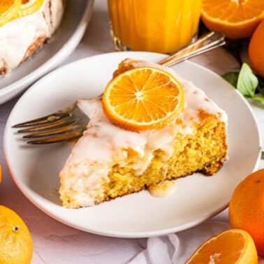 A slice of glazed orange clementine cake garnished with an orange slice is served on a white plate, surrounded by whole and halved oranges, with a glass of orange juice in the background.
