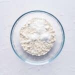 A clear glass bowl containing a mixture of white flour and baking powder on a light textured surface.