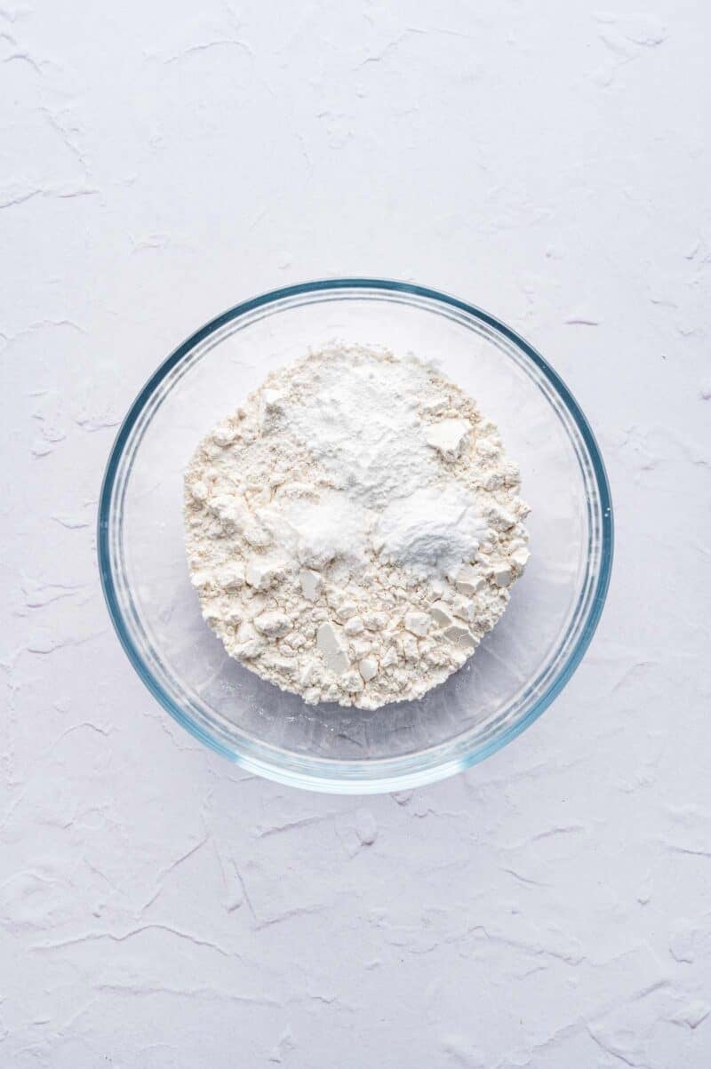 A clear glass bowl containing a mixture of white flour and baking powder on a light textured surface.
