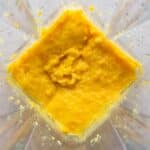 A close-up view of a blender containing a thick, yellow-orange mixture with some chunky texture.