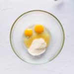 A glass bowl containing two whole eggs, sugar, and liquid on a white surface, with a metal whisk partially visible in the corner.