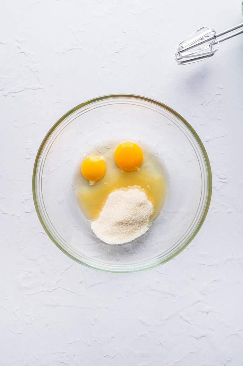 A glass bowl containing two whole eggs, sugar, and liquid on a white surface, with a metal whisk partially visible in the corner.
