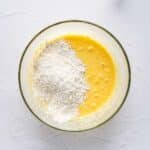 A glass bowl with a yellow batter mixture and a mound of flour on top, with a hand mixer and batter residue visible nearby.