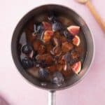 A saucepan containing chopped figs immersed in liquid, placed on a light pink surface with a wooden spoon nearby.