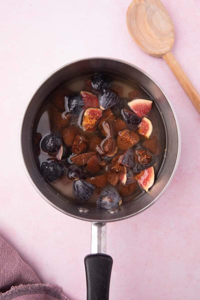 A saucepan containing chopped figs immersed in liquid, placed on a light pink surface with a wooden spoon nearby.