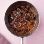 A saucepan filled with a dark, chunky mixture, possibly a simmering chutney or sauce, sits on a light pink surface next to a folded purple cloth.