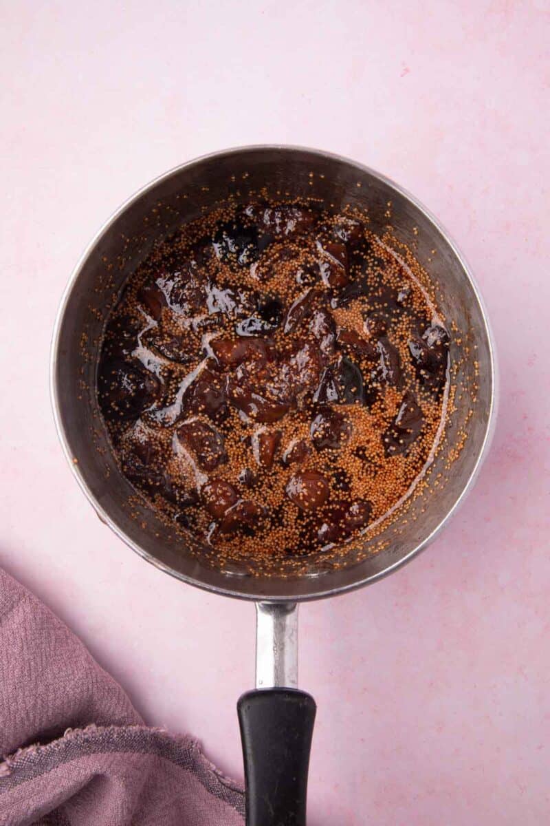 A saucepan filled with a dark, chunky mixture, possibly a simmering chutney or sauce, sits on a light pink surface next to a folded purple cloth.