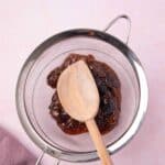 A wooden spoon resting on fig jam in a metal strainer over a pink surface with a mauve cloth nearby.