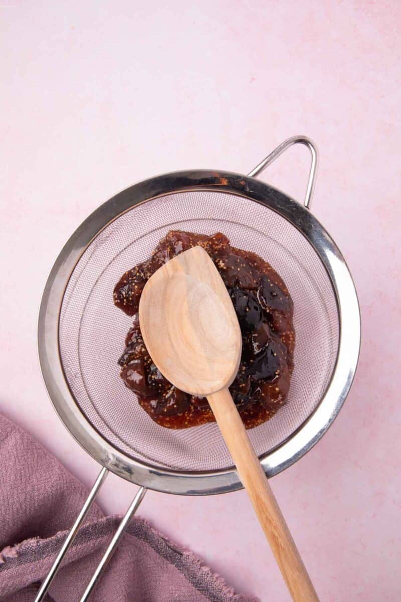 A wooden spoon resting on fig jam in a metal strainer over a pink surface with a mauve cloth nearby.