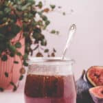 A glass jar filled with fig syrup and a spoon inside, placed on a light surface with a potted plant and fresh figs in the background.