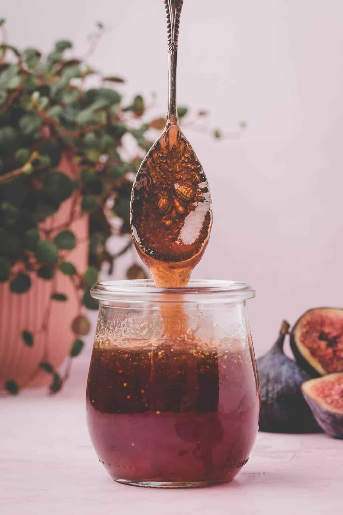 A spoon dripping fig syrup into a glass jar, with fresh figs and a potted plant in the background.