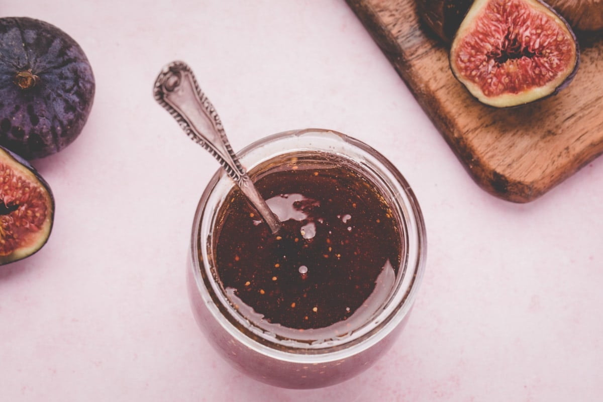 A jar of fig syrup with a spoon inside, surrounded by fresh figs and fig halves on a wooden board, on a light pink surface.