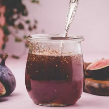A glass jar of fig syrup with a spoon inside, next to fresh figs and a wooden board, set against a pink background.