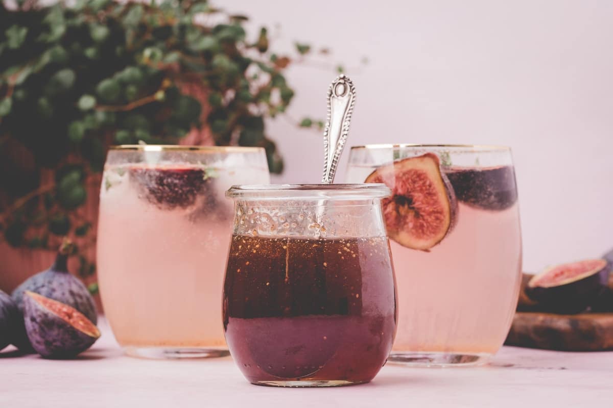 A jar of fig syrup with a spoon in it is placed in front of two glasses of pink drinks garnished with fig slices. Figs and a plant are in the background.