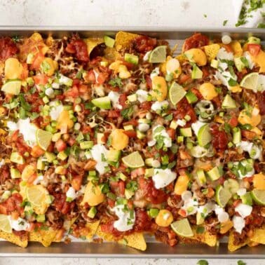 A sheet pan filled with loaded nachos topped with cheese, sour cream, diced tomatoes, avocado, jalapeños, green onions, salsa, and lime wedges.