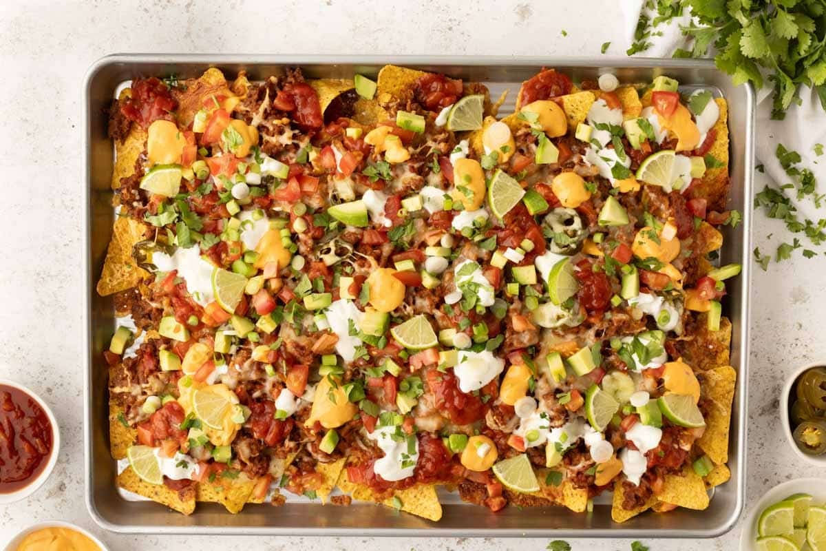 A sheet pan filled with loaded nachos topped with cheese, sour cream, diced tomatoes, avocado, jalapeños, green onions, salsa, and lime wedges.