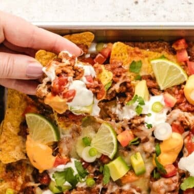 A hand holding a loaded nacho chip above a tray of sheet pan nachos topped with ground meat, melted cheese, sour cream, diced tomatoes, avocado, lime wedges, and green onions.