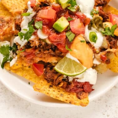 Plate of ground beef nachos topped with ground meat, chopped tomatoes, avocado, salsa, sour cream, cilantro, and a lime wedge.